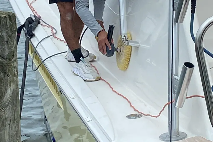 A Platinum Prestige detailer in a branded shirt working a rotary polisher along the hull side of a sport fisherman from the dock