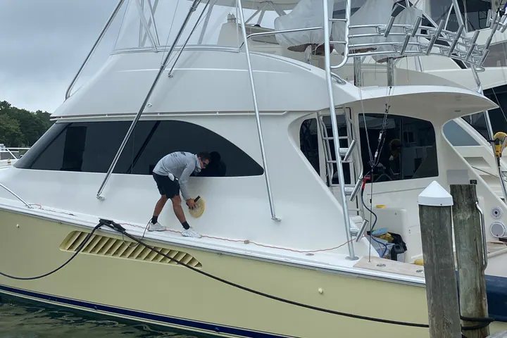 A detailer working on the side deck of a cream-hulled sport fisherman at the dock, wash gear laid out around him